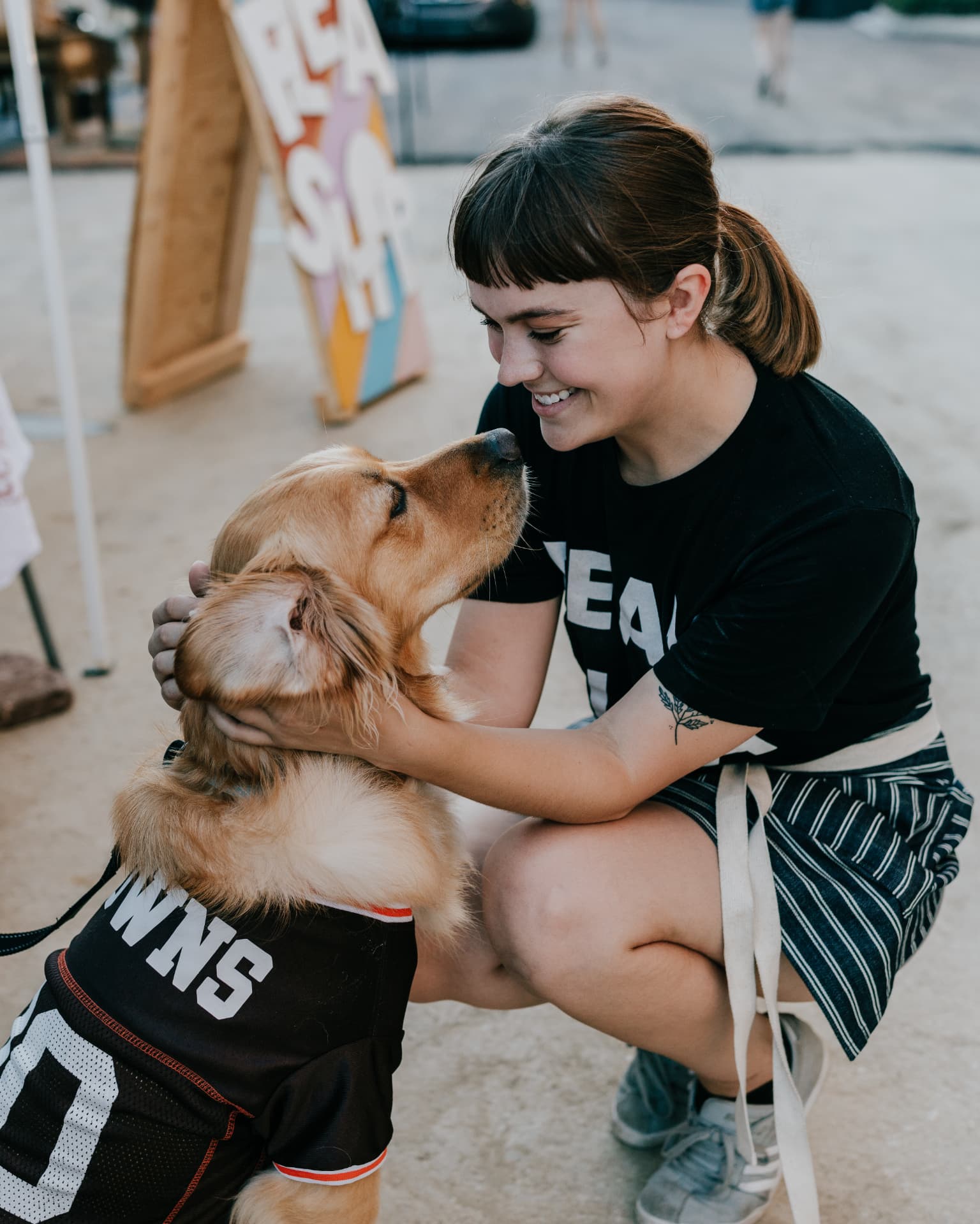 Smiling woman kneeling to greet a golden dog in a jersey at an outdoor flea market